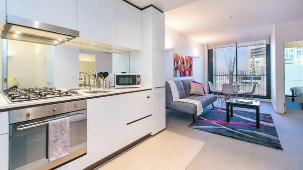 A modern kitchen with a gas stove, microwave, and oven, a gray sofa, and a large window with a view of buildings.