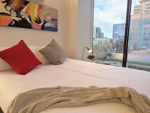 A modern hotel room with a large bed, a city view, a painting on the wall, and a lamp on the nightstand.