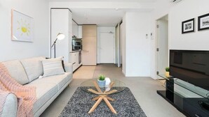Ein modernes Wohnzimmer mit einer Couch, einem Glastisch, einem Fernseher und einer Küche im Hintergrund.