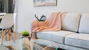 Ein modernes Wohnzimmer mit grauem Sofa, einem Glastisch, einem weißen Sessel, einer Strickdecke und einer Zierpflanze.