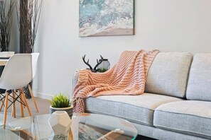 Un salon moderne avec un canapé gris, une table basse en verre, une chaise blanche, une petite plante et un tableau abstrait accroché au mur.