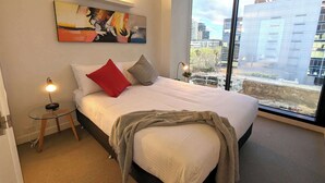 Ein Hotelzimmer mit einem großen Bett, einem Gemälde an der Wand, Blick auf Gebäude durch das Fenster und einem kleinen Nachttisch mit einer Lampe.