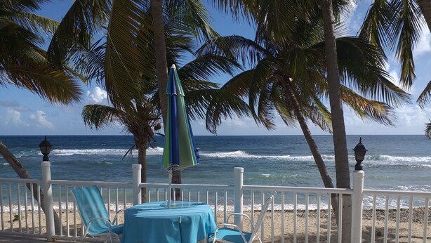 Een terras aan het strand met een tafel en stoelen, palmbomen en uitzicht op de oceaan.