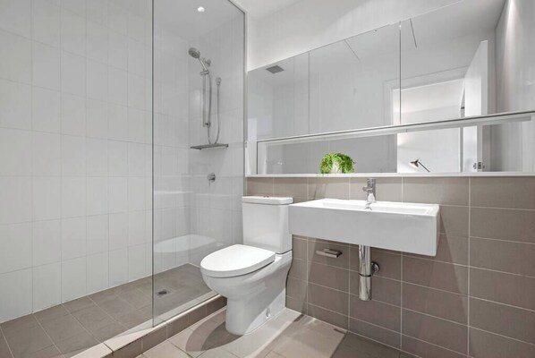 Une salle de bain moderne avec une douche à parois de verre, des toilettes blanches et un lavabo surmonté d’un miroir.