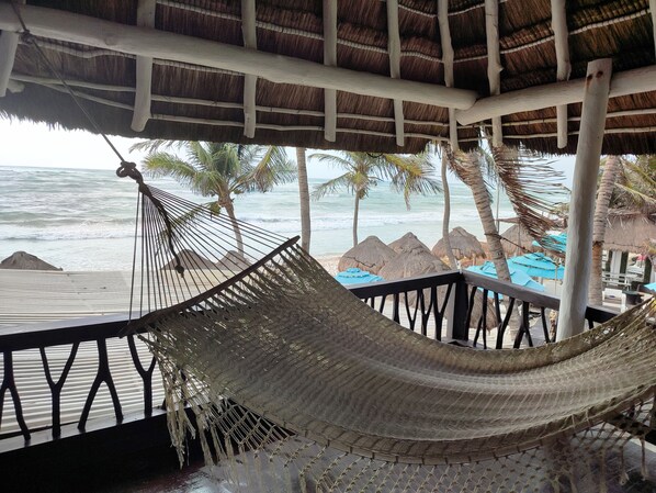 A hammock under a thatched roof, overlooking a beach with palm trees and thatched umbrellas. - Villa Las Estrellas Tulum - located at the party zone (Tulum)