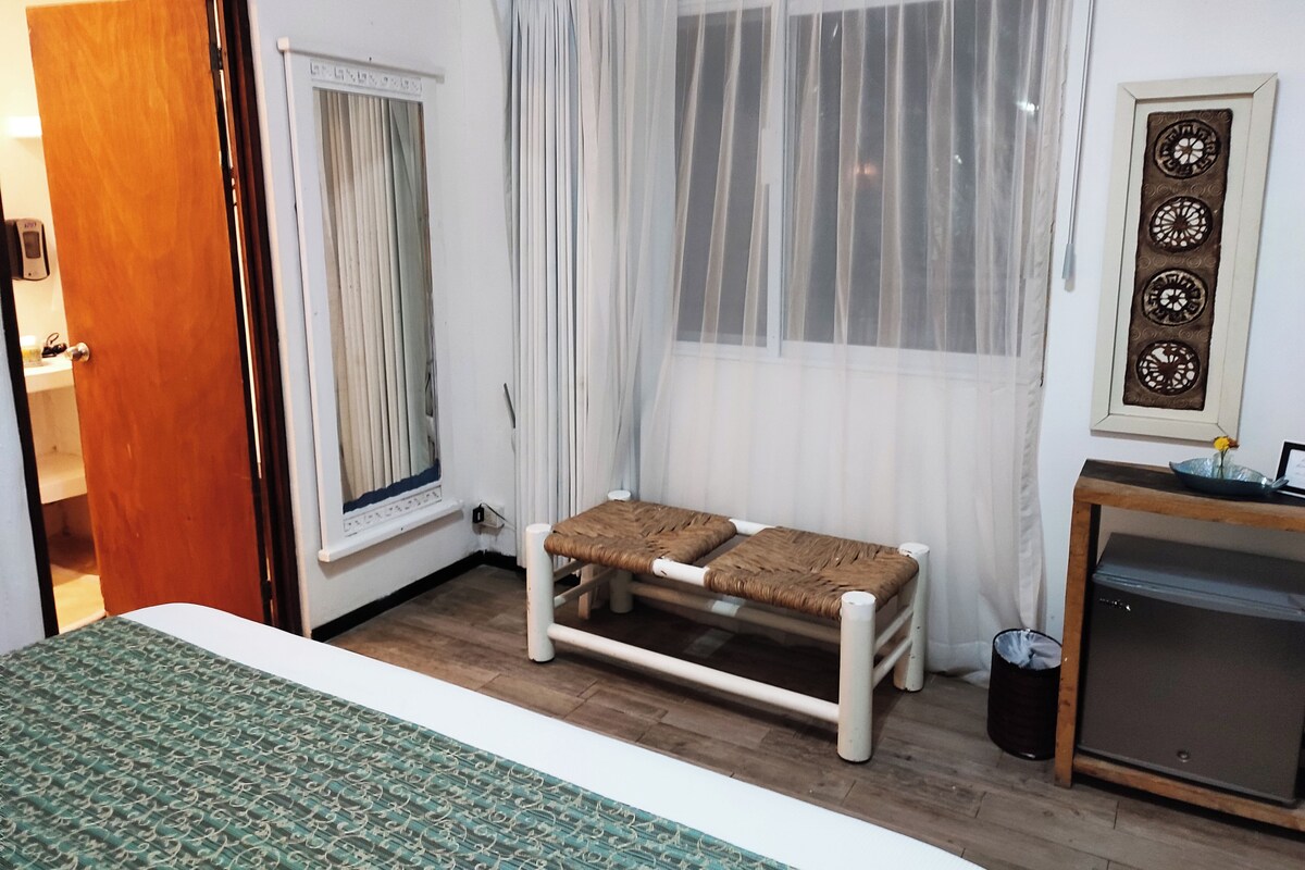 Una habitación con cama, un banco, una ventana con cortinas translúcidas, un adorno en la pared y una mesita con un cuenco y una flor.
