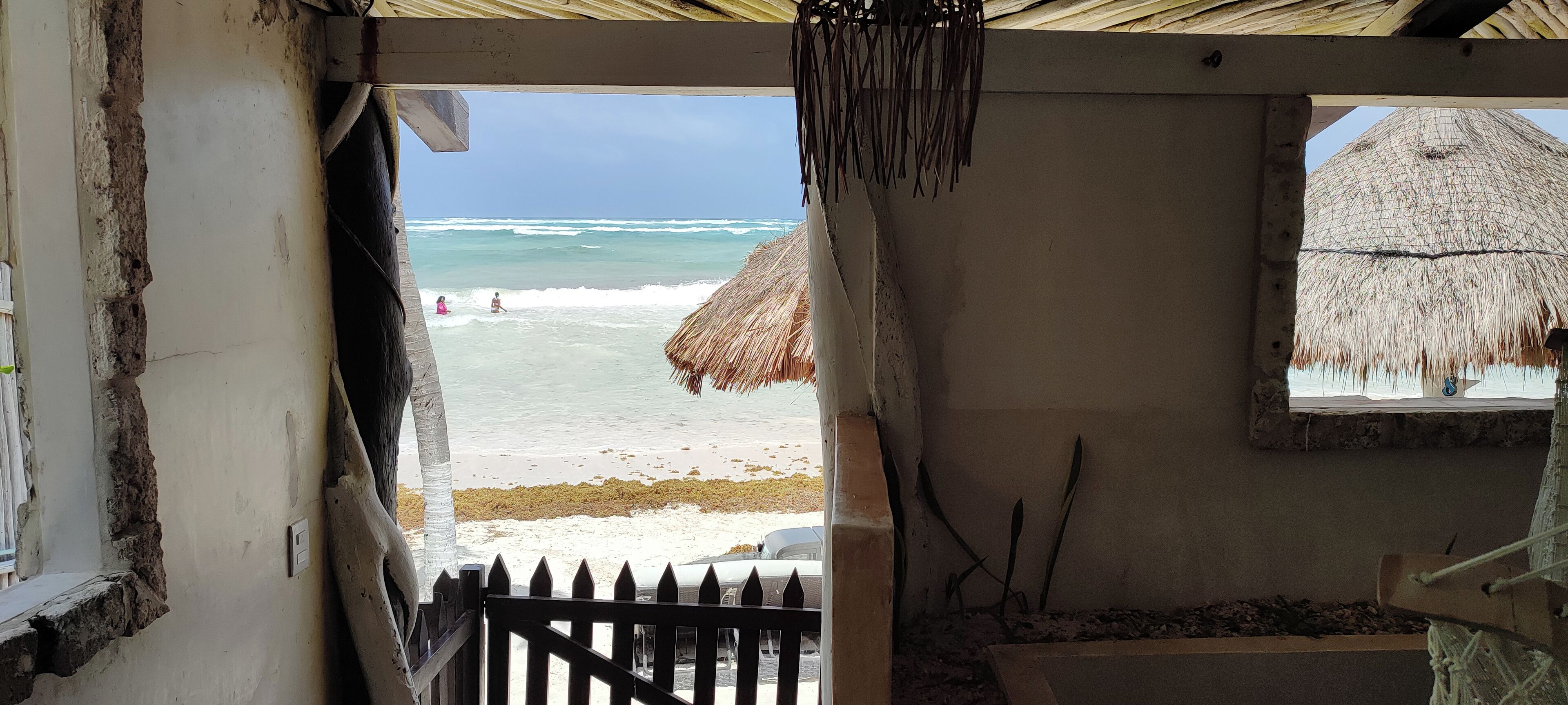 Blick auf einen Strand von einer überdachten Veranda aus, mit einem Reetdach, einem Holzzaun und einem Fenster mit Meerblick.
