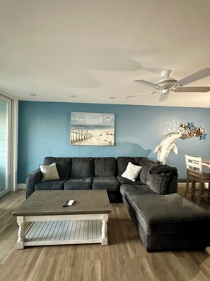 A living room with a sectional sofa, a coffee table, a ceiling fan, and a painting of a beach scene.