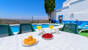 A table with croissants, cherries, and two glasses of orange juice by a pool with blue chairs and a white building in the background. - Sierra Tejeda  -  El Lagarillo Alcaucín by Ruralidays (Málaga)