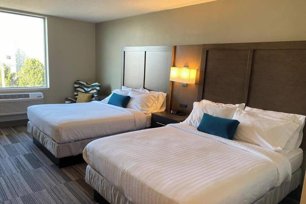 A hotel room with two beds, a chair, a lamp, and a window.