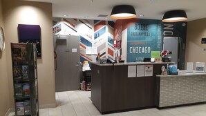 A hotel reception area with a counter displaying city names and a directory. There is a magazine rack to the left and a door leading to another room. - Simple Suites Boise Airport (Boise)