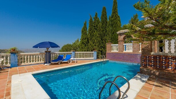 Una piscina con acqua blu, circondata da un solarium piastrellato, lettini blu e un ombrellone azzurro.