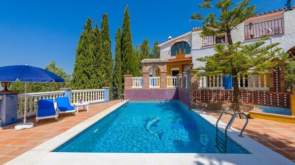 A swimming pool with a tiled deck, lounge chairs, and a house with a tiled facade and balcony.