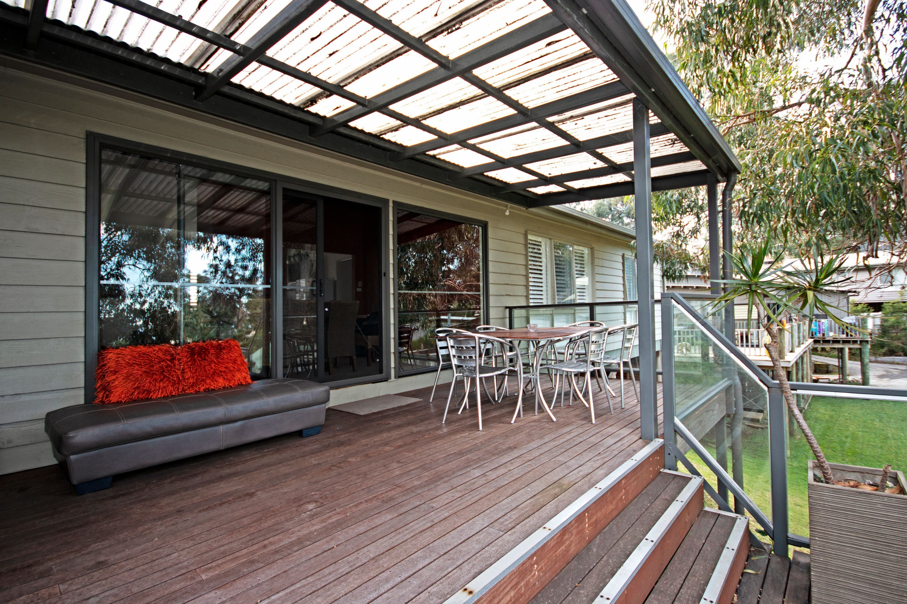 A covered outdoor area with a wooden deck, a seating area with a red cushion, and a table with chairs.
