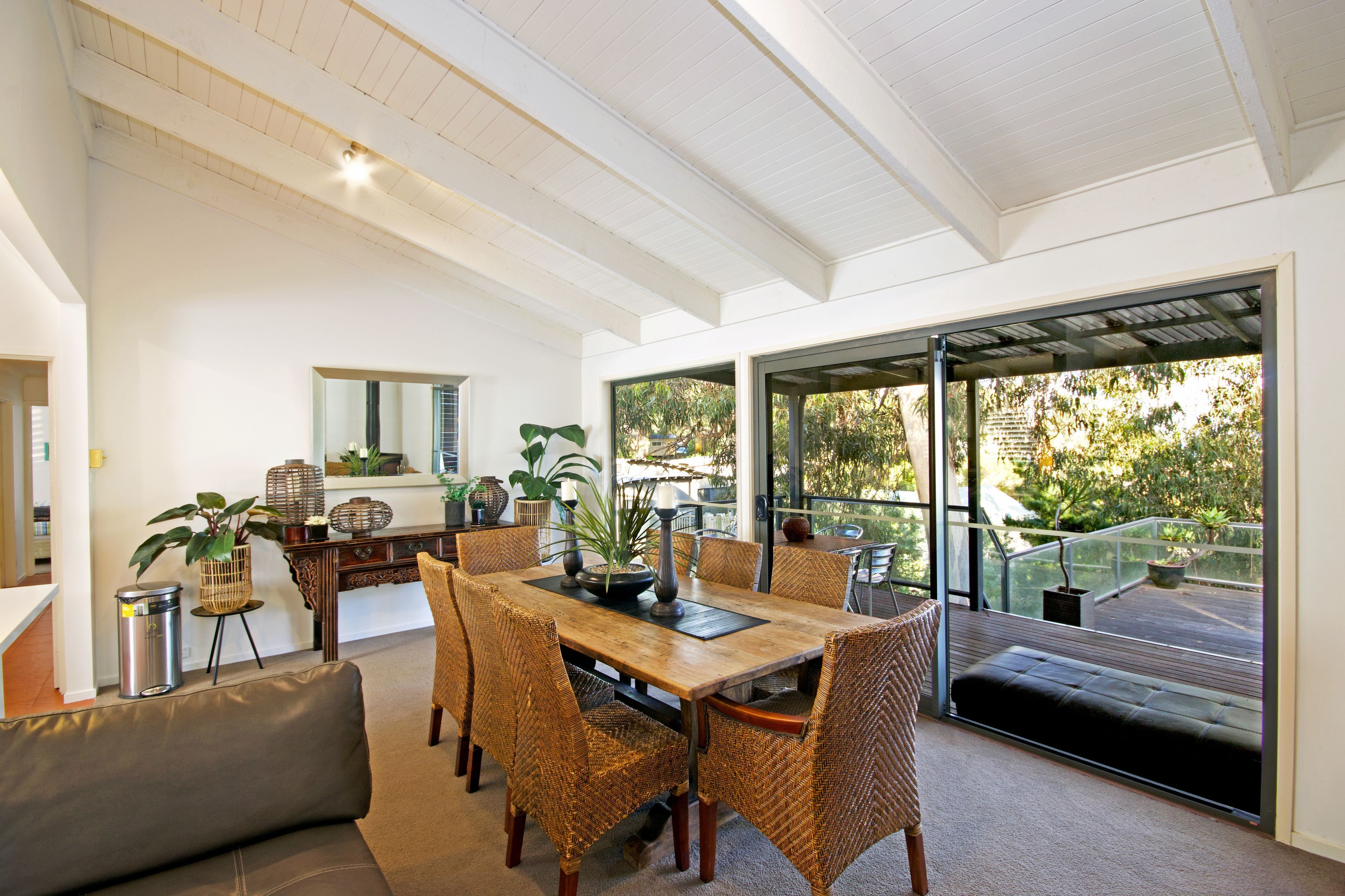 Uma sala de jantar ampla com mesa de madeira e cadeiras de vime, uma mesinha lateral com objetos decorativos, e vista para o exterior através de grandes portas de vidro.