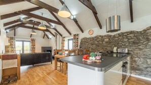 A modern kitchen with a central island, wooden beams, stone wall, and a dining area with chairs and a table.