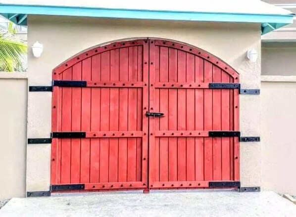 Une porte rouge à deux battants, agrémentée de détails noirs, encadrée dans un mur de couleur beige surmonté d’un toit bleu.