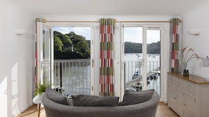 A modern living room with a grey sofa, a view of a lake with boats, and a balcony with striped curtains. - This holiday home is situated right on the water’s edge within the Malpas Marina (Malpas)