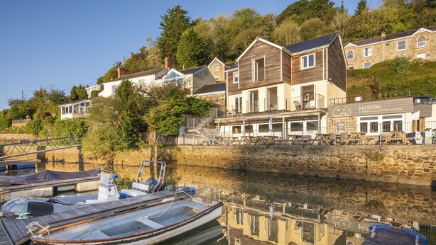 A waterfront property with a wooden boat docked, a coffee shop named 'Heron Farm & Coffee Shop', and residential buildings in the background. - This holiday home is situated right on the water’s edge within the Malpas Marina (Malpas)