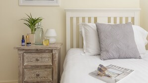 Ein ordentlich bezogenes Bett mit weißem Kopfteil, ein Nachttisch mit Pflanze, Lampe und Zeitschrift.