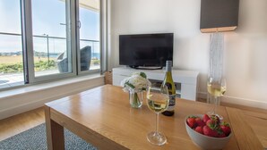 A room with a large window, a flat-screen TV, a wooden table with a wine bottle, glasses, and a bowl of strawberries. - A stunning first floor apartment within an award-winning development at Porth. (Newquay)