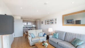 A modern living room with a sofa, a coffee table with drinks, and a kitchen in the background. - A stunning first floor apartment within an award-winning development at Porth. (Newquay)
