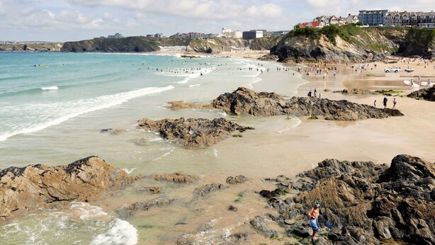 A coastal beach with rocky areas, a sandy shore, and buildings in the background. - A stunning first floor apartment within an award-winning development at Porth. (Newquay)