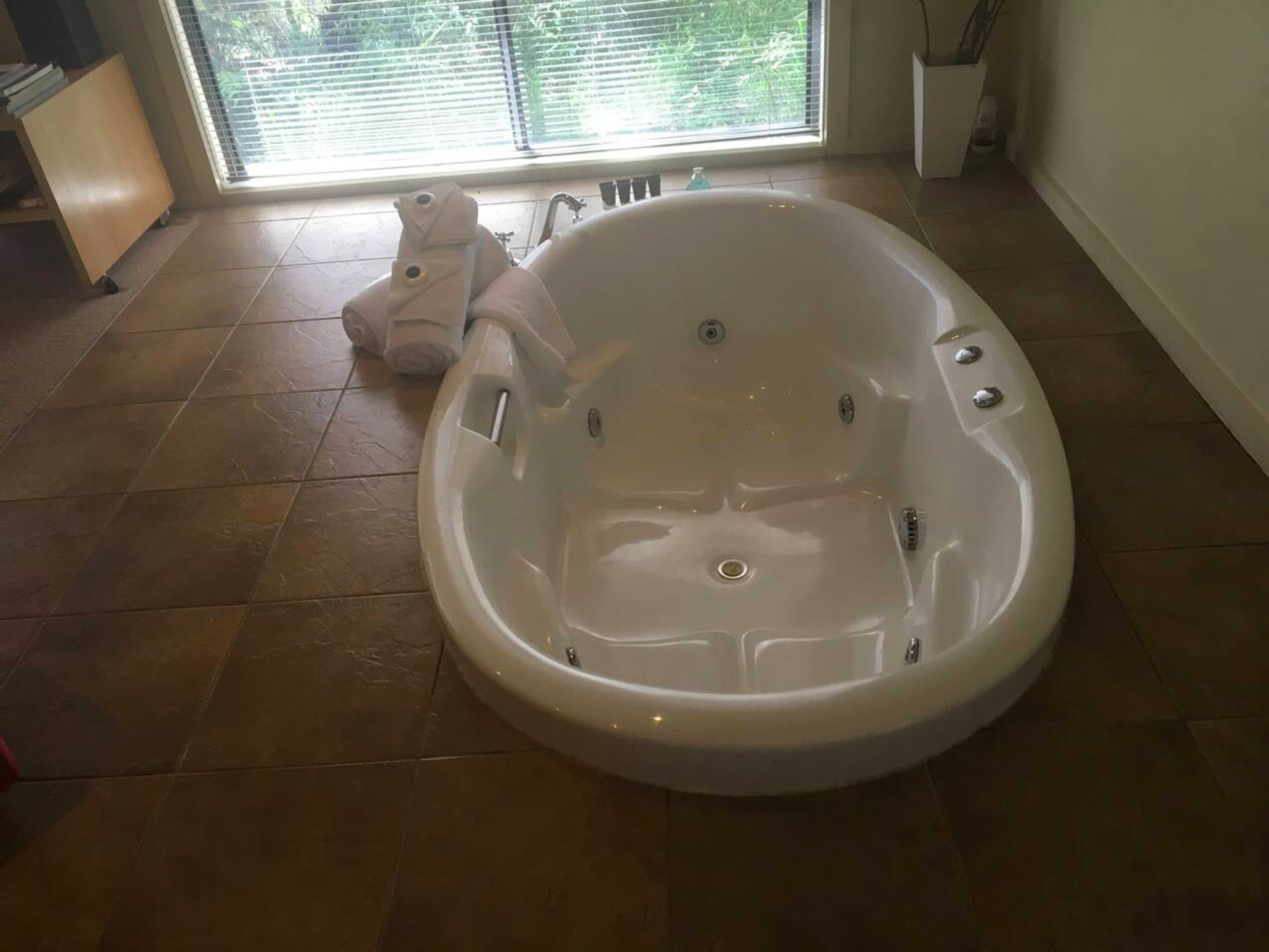 A large, empty bathtub in a room with a tiled floor and a window with blinds.