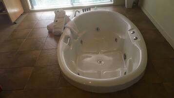 A large, empty bathtub in a room with a tiled floor and a window with blinds.