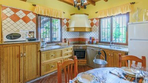 A kitchen with wooden cabinets, a microwave, a stove, and a dining area with a table set for two. - Las Lomas de San Rafael Periana by Ruralidays (Málaga)