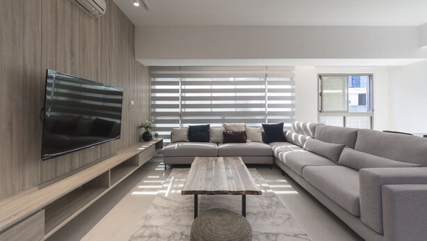 Una sala de estar moderna con un televisor de pantalla plana grande montado en una pared de madera, un sofá seccional gris, una mesa de centro de madera y un ventanal con persianas.