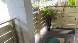 Une terrasse en bois avec un paillasson aux motifs verts et bleus, une clôture en bois et un pot de fleurs.