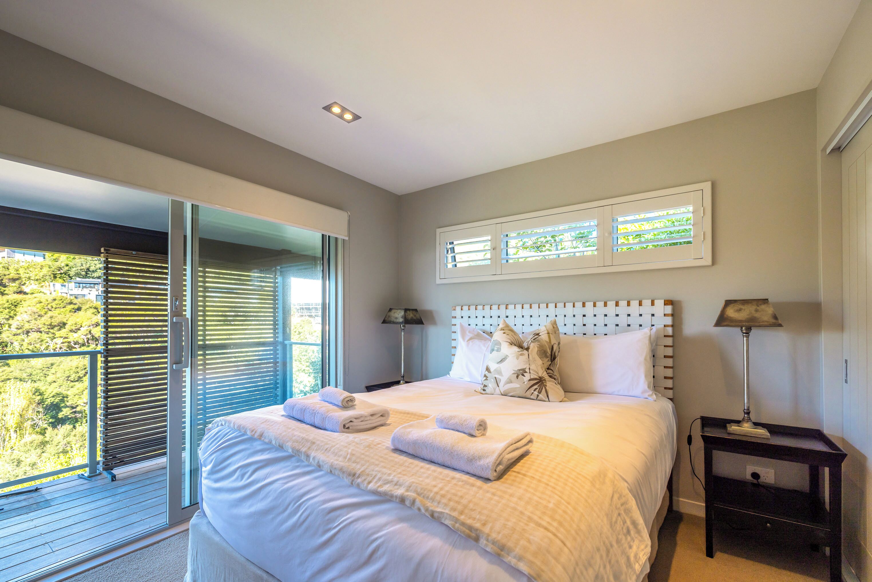 A bedroom with a large bed, two bedside lamps, a window with blinds, and a view of greenery outside.