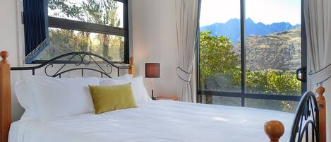 A bedroom with a bed, a nightstand, a lamp, and a view of mountains through large windows.