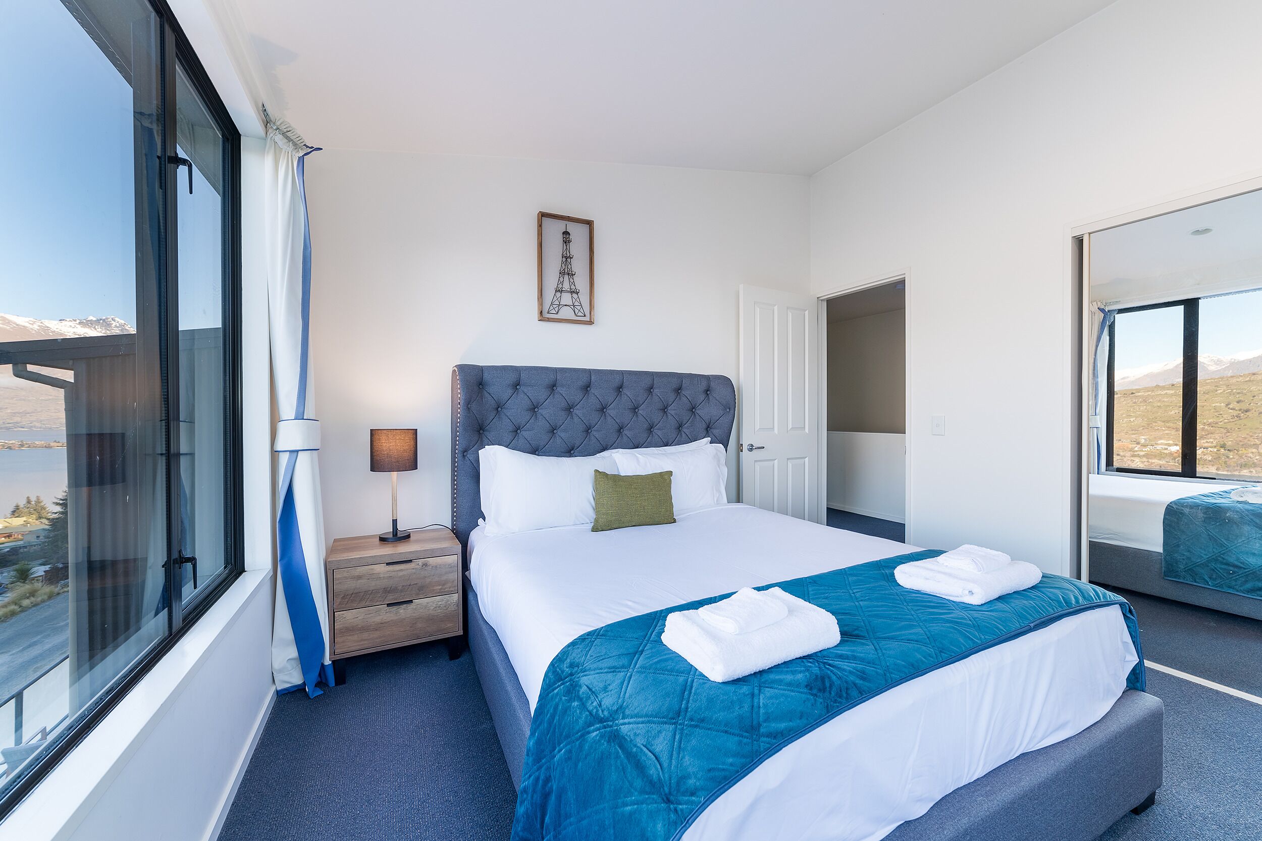 A hotel room with a large bed, a nightstand, a lamp, a framed picture on the wall, and a view of the outdoors.