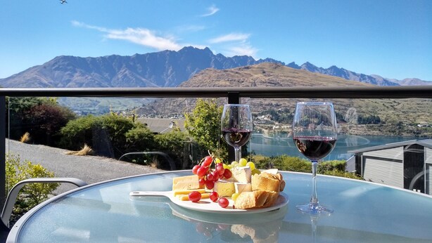 Una mesa con un plato de queso, uvas y pan, dos copas de vino y una vista panorámica de la montaña.