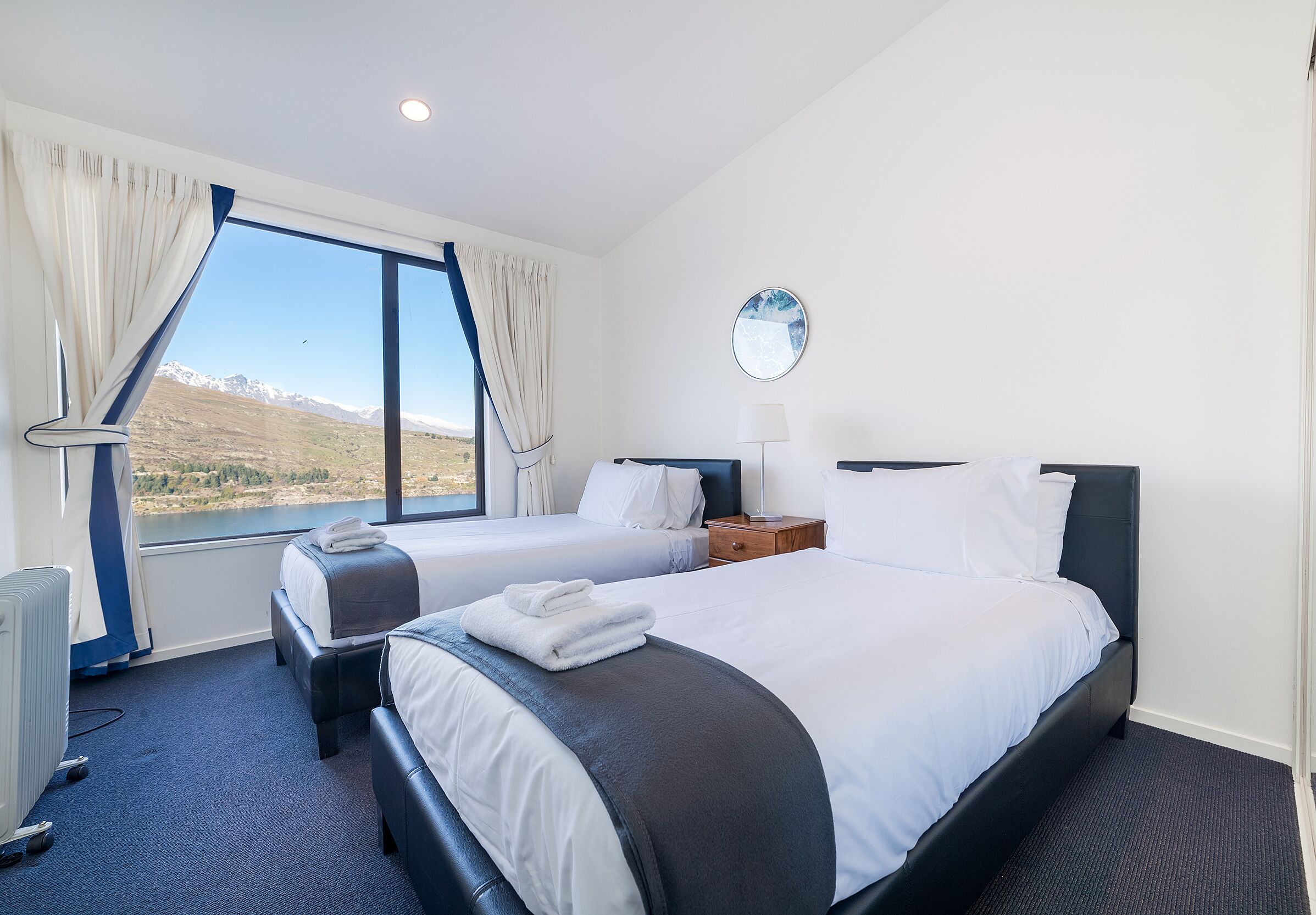 A hotel room with two beds, a large window with a view of mountains, a round mirror, and a bedside table with a lamp.