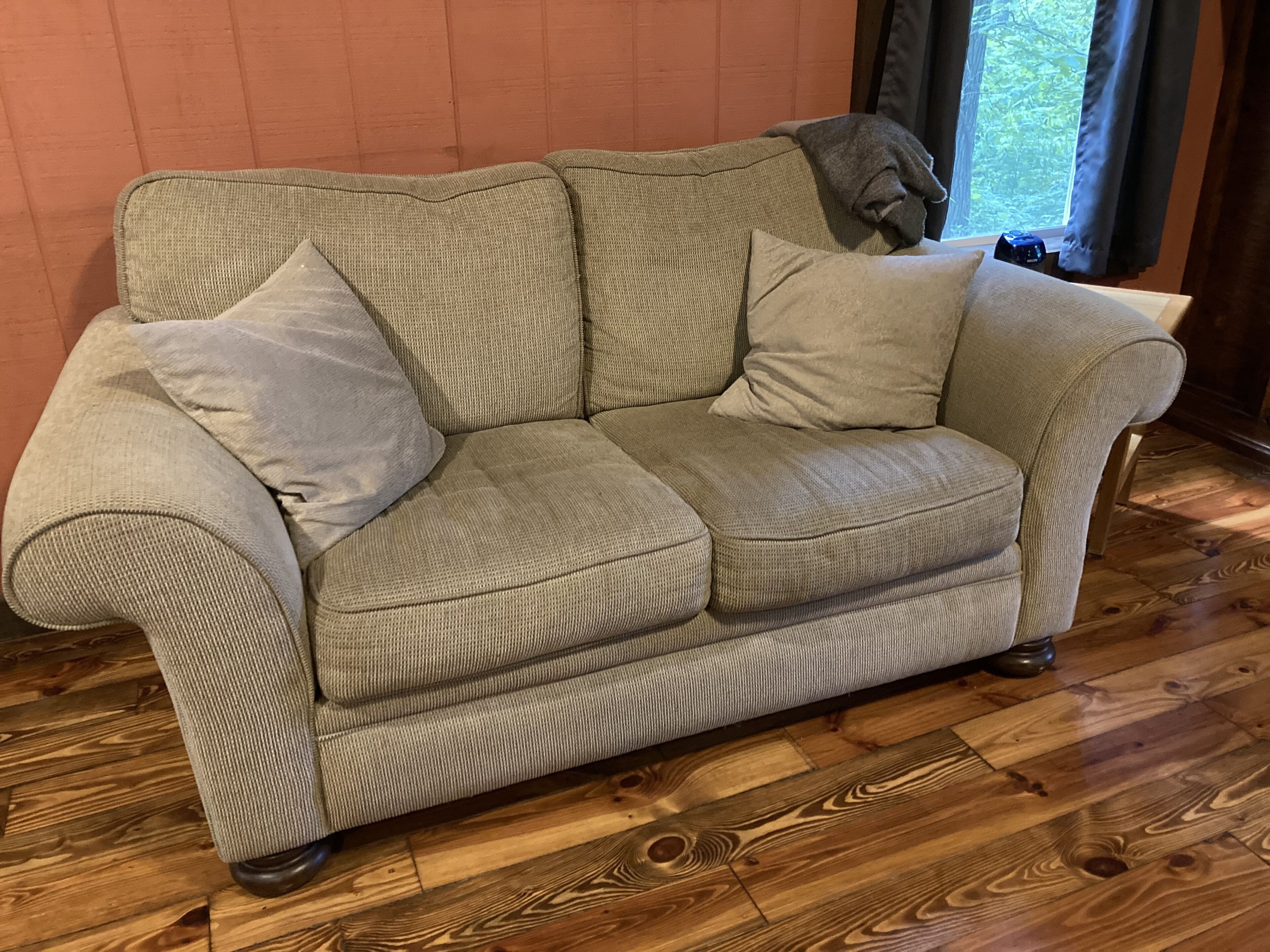 A beige sofa with two pillows, positioned against a wooden floor and a plain wall.