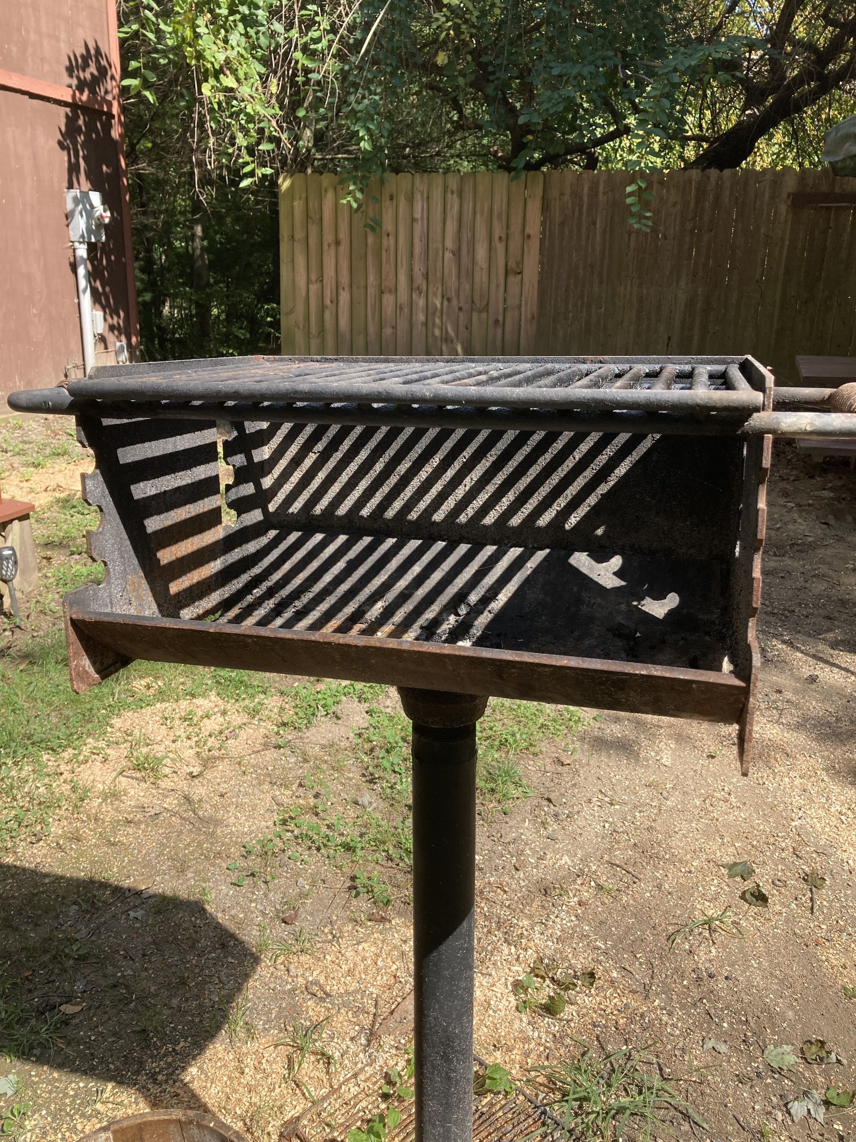 Una parrilla metálica de diseño con listones, sostenida por un único poste vertical, ubicada en un patio con cerco de madera y follaje al fondo.