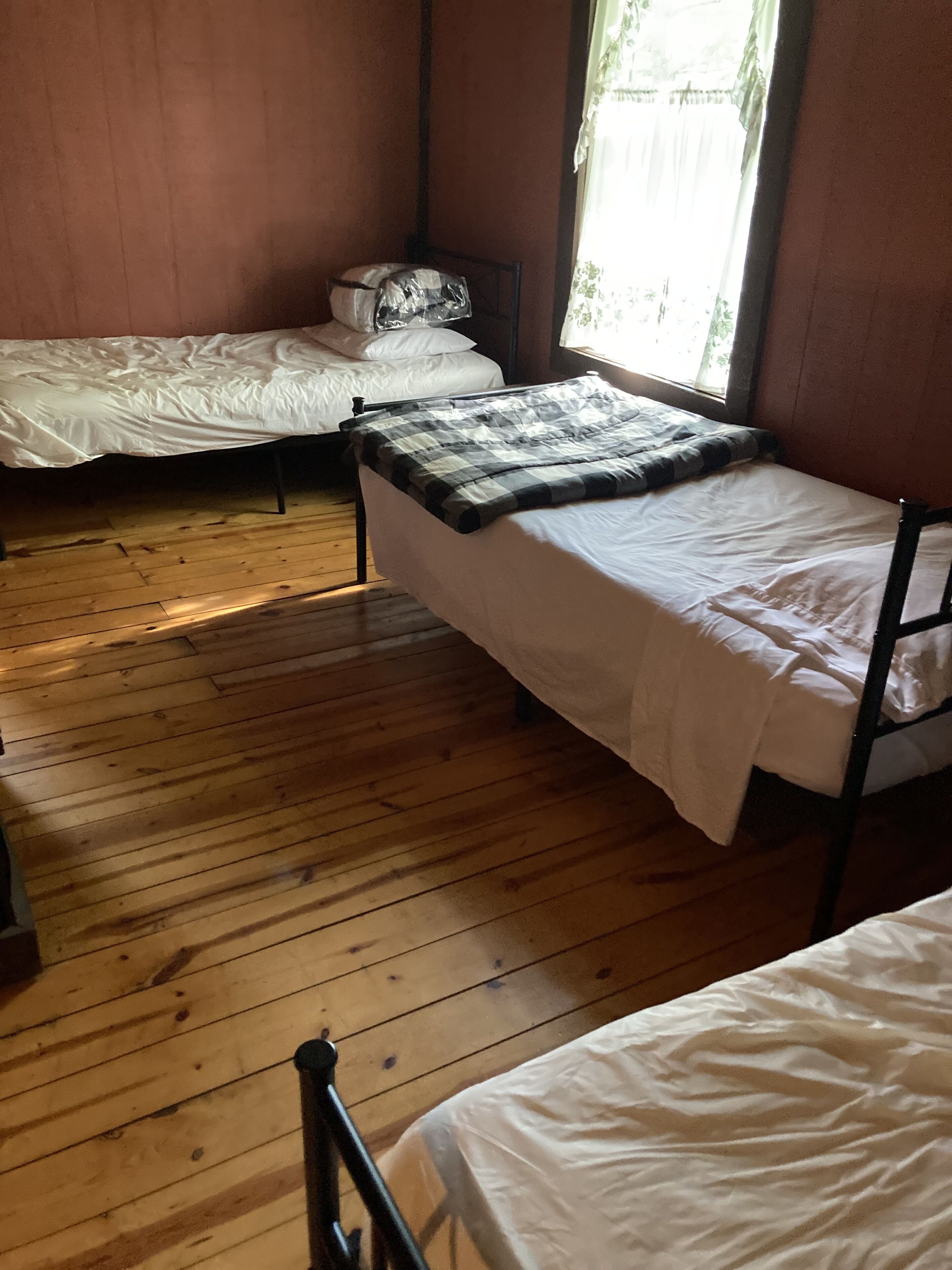 A dormitory room with two single beds, wooden flooring, and a window with curtains.