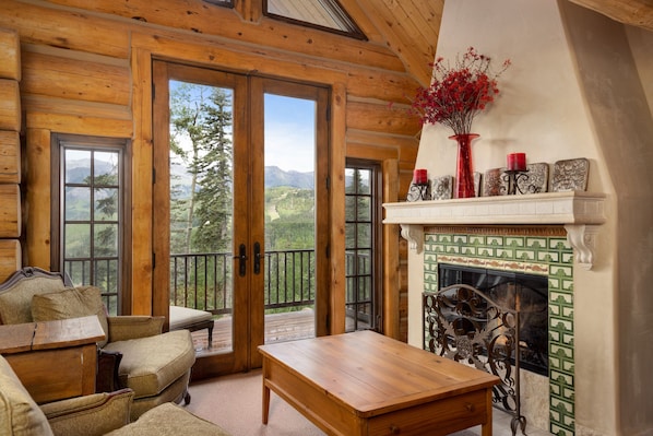 A log cabin interior with a fireplace, wooden furniture, and a balcony with a mountain view.