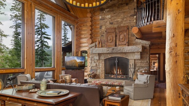 A log cabin interior with a stone fireplace, wooden furniture, and large windows overlooking a forest. - HILLTOP HIDEAWAY- A Magical Mountain Ski Estate with Game Room, Hot Tub, and Vie (Telluride)