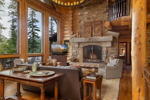 A log cabin interior with a stone fireplace, wooden furniture, and large windows overlooking a forest.
