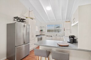 A modern kitchen with a stainless steel refrigerator, a gas stove, and a built-in oven. There are two grey chairs at the island and a window with vertical blinds.