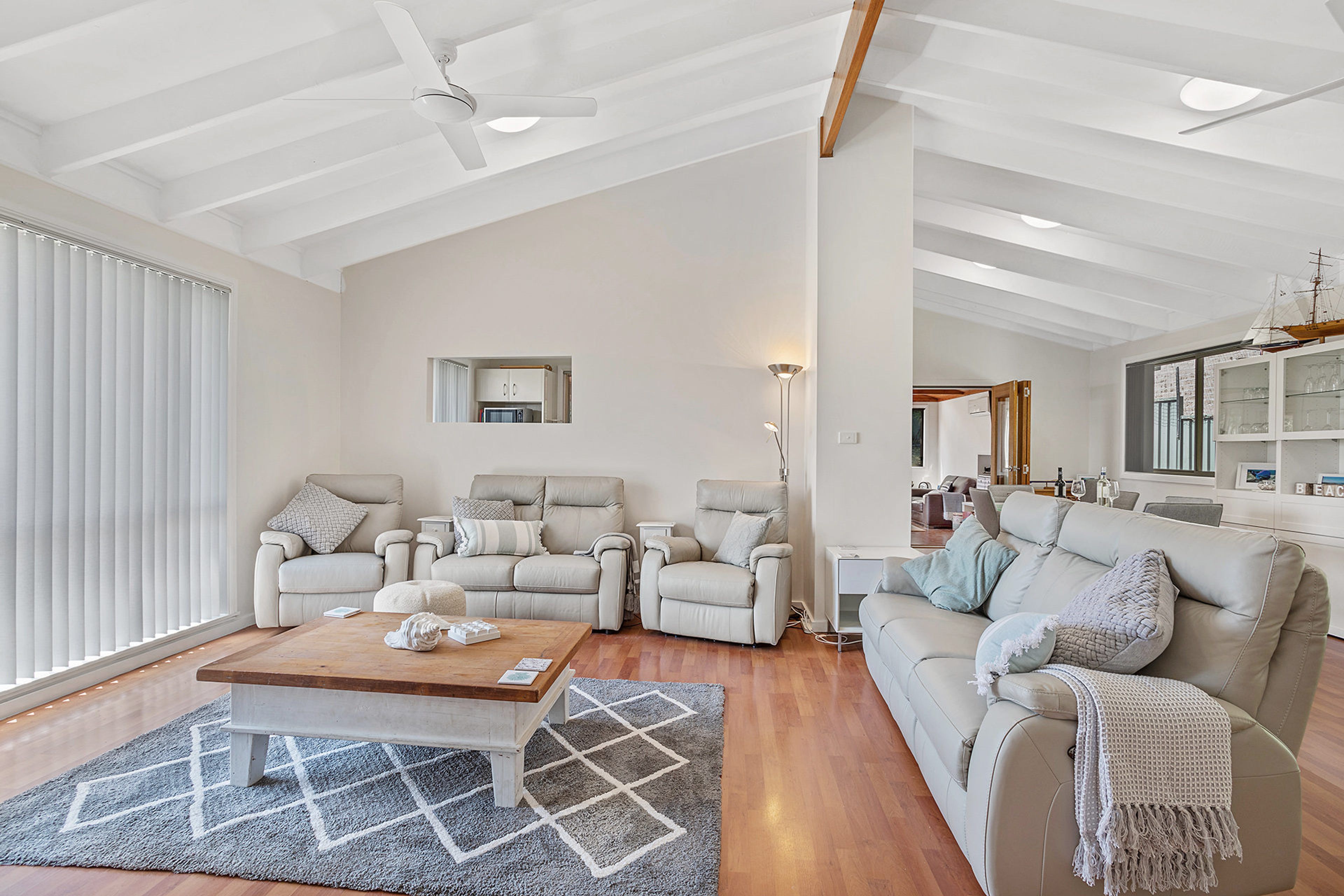 A spacious living room with a high ceiling, a ceiling fan, and a large window with blinds. There is a seating area with two sofas and a coffee table.
