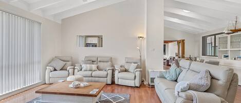 Ein geräumiges Wohnzimmer mit hoher Decke, Deckenventilator und einem großen Fenster mit Jalousien. Es gibt eine Sitzecke mit zwei Sofas und einem Couchtisch.