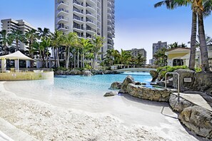 A resort pool area with clear water, surrounded by palm trees and a sandy beach.