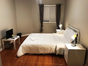 A neatly arranged bedroom with a bed, bedside tables, a desk with a computer, and a window with curtains. - Guardian Cottage (Mississauga)