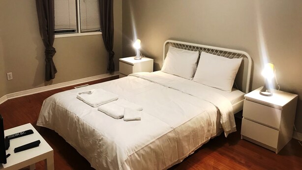 A neatly made bed with white linens and two bedside lamps, a small table with a remote control, and a window with curtains. - Guardian Cottage (Mississauga)