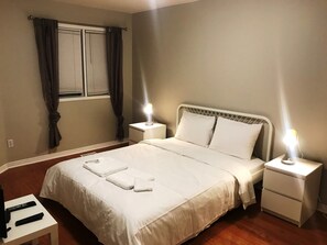 A neatly made bed with white linens and two bedside lamps, a small table with a remote control, and a window with curtains. - Guardian Cottage (Mississauga)